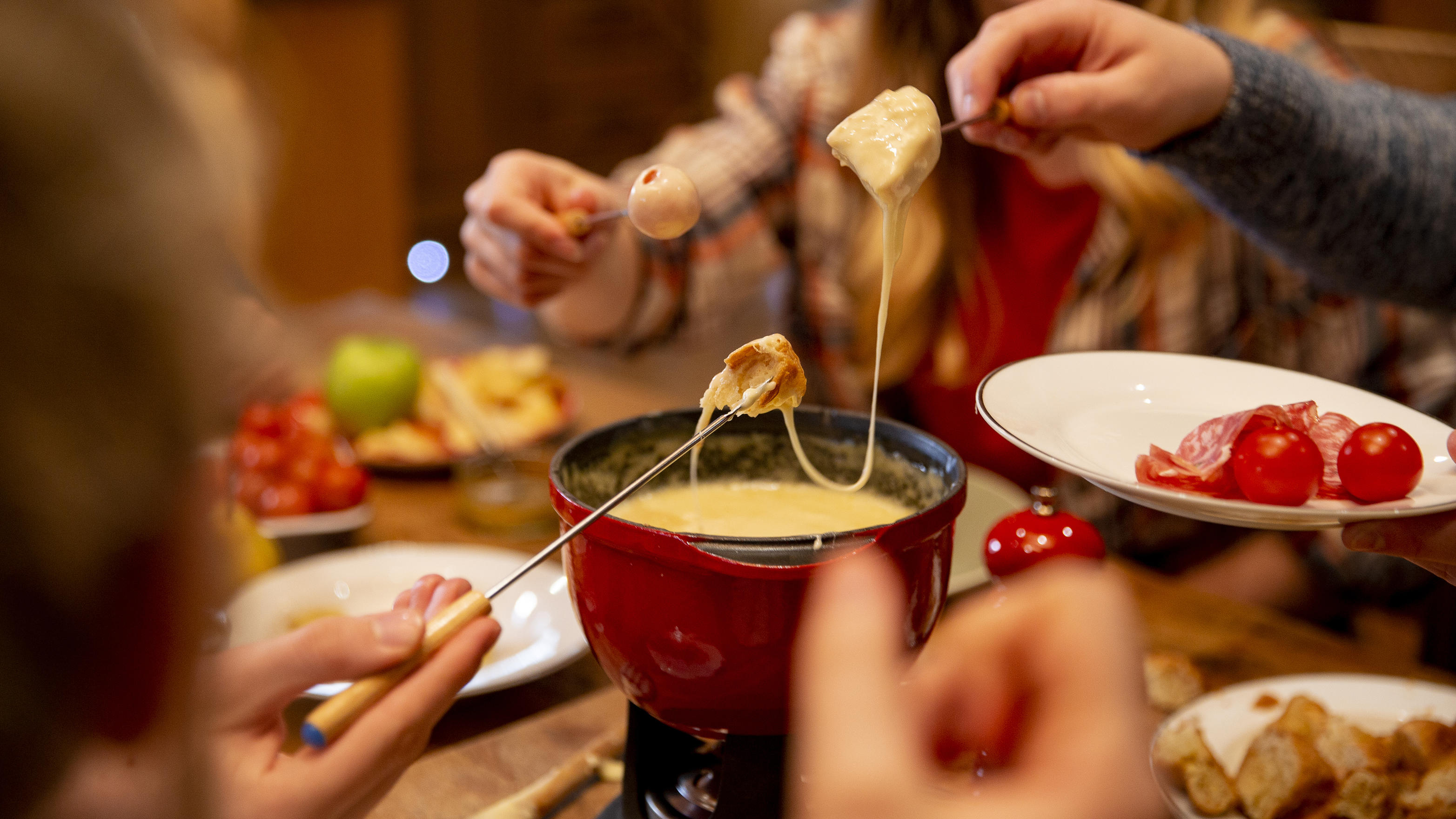 Geschmolzener Käse in einem roten Topf auf einem Küchentisch und vielen Händen drumherum.
